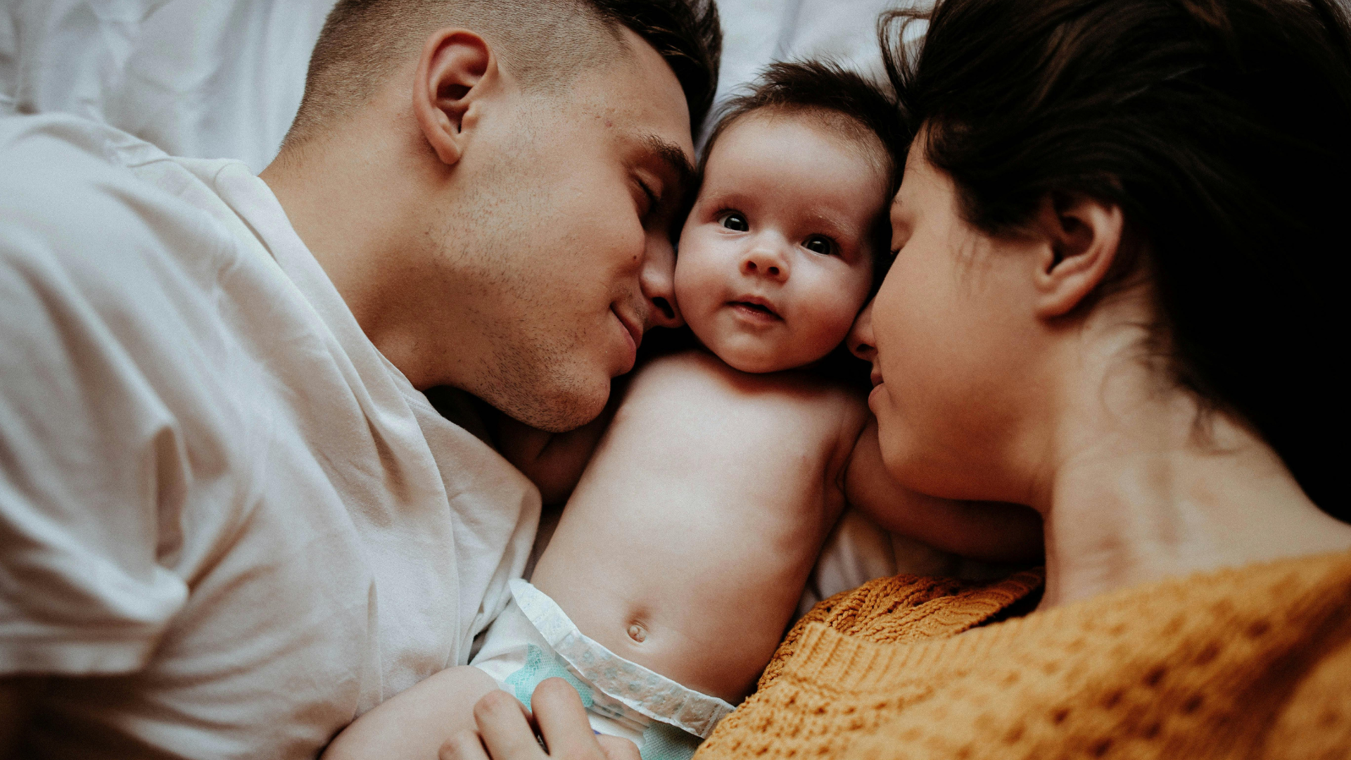  Un couple allongé sur un lit entoure tendrement un bébé. L'homme à gauche touche doucement le bébé avec son visage, tandis que la femme à droite le regarde affectueusement. Le bébé, en couche, regarde l'appareil photo avec curiosité et un léger sourire.